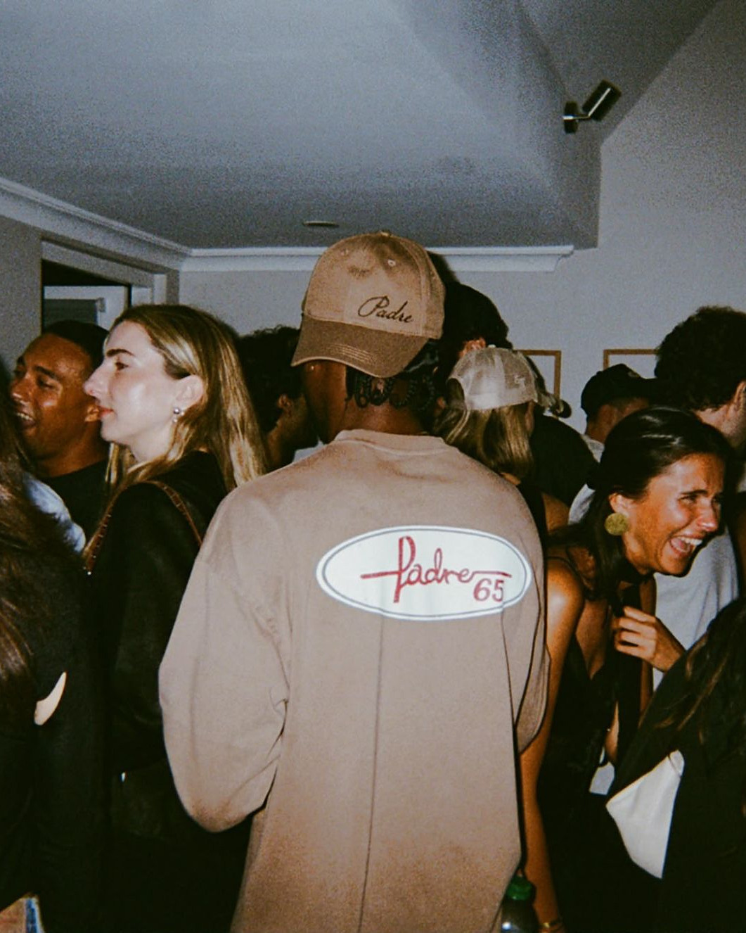 Guests at the last Padre65 party; at the centre, a man seen from behind in a Padre cap and a taupe top with the Padre65 script on the back.