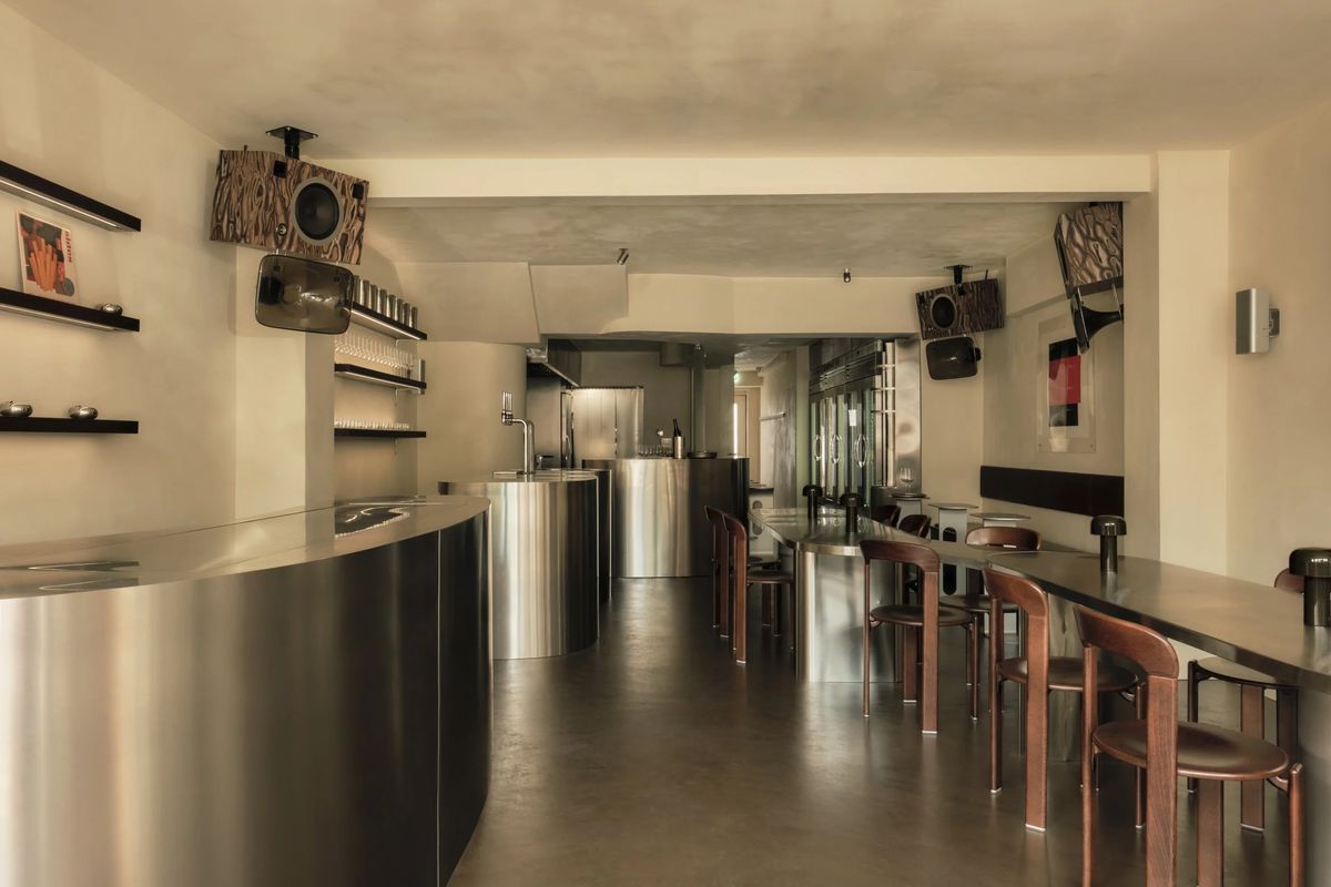Inside Kiez: a long curved stainless-steel counter, wooden bar stools along a steel bar, and wood-veneer speakers mounted on the walls.